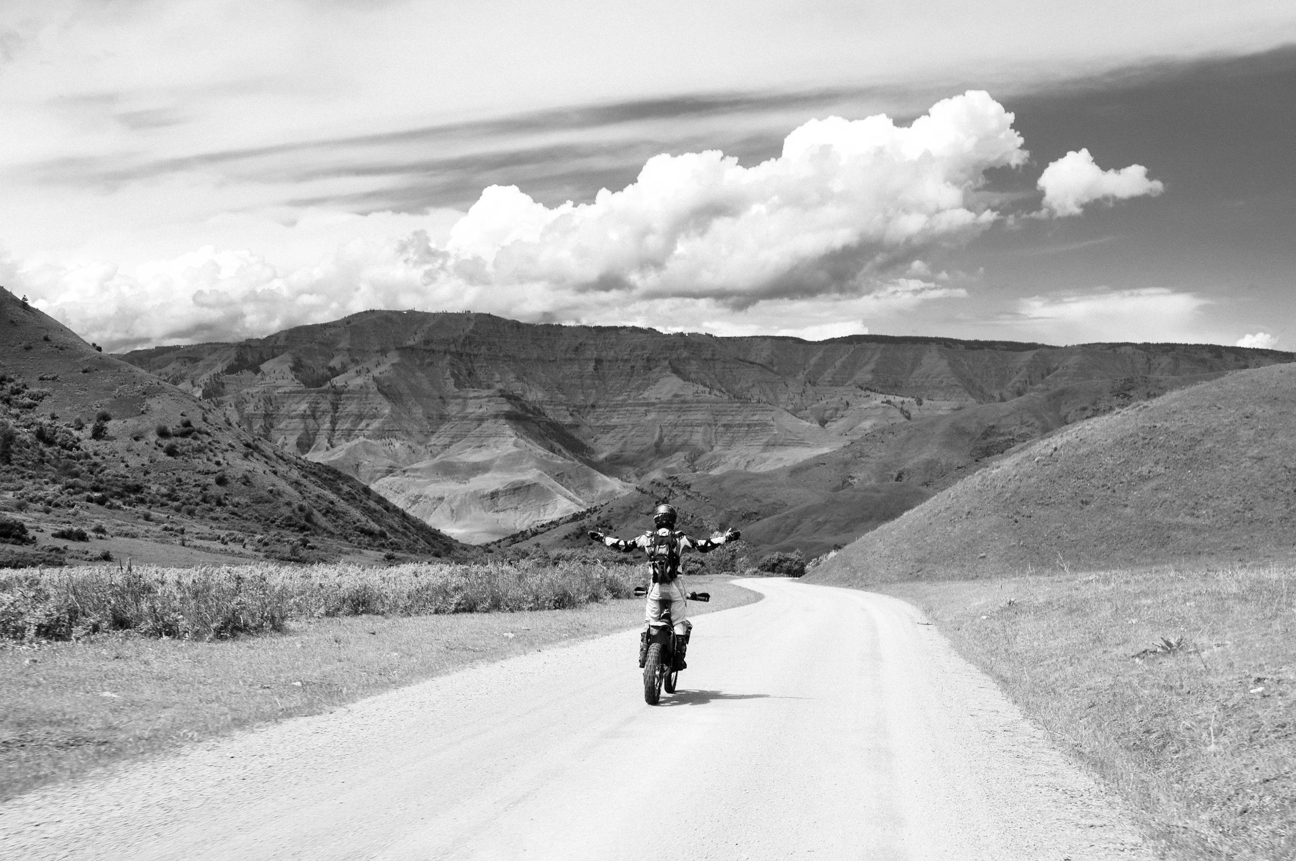 Black and white back view of anonymous biker with outstretched arms riding motorbike on curved roadway between mounts under cloudy sky